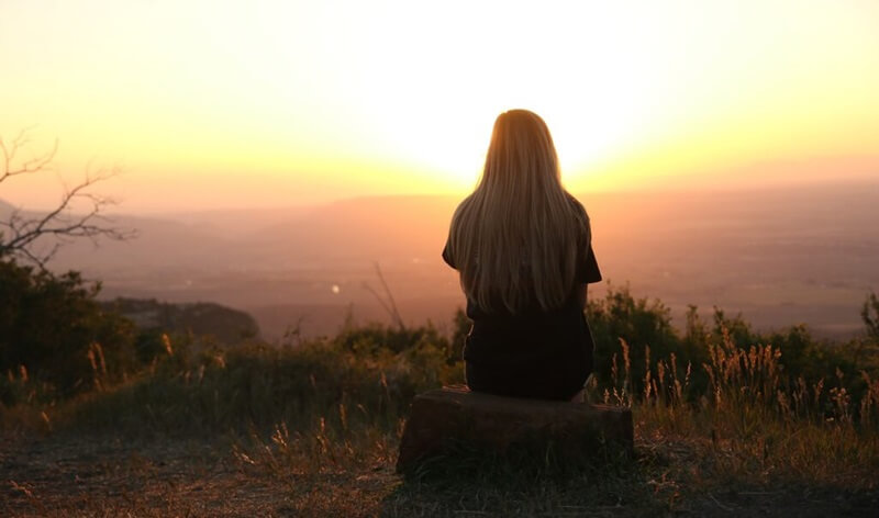 野原に座って夕日を眺めがめている女性